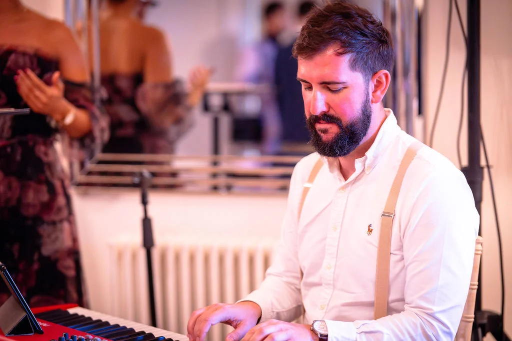 A bearded pianist wearing a white shirt and braces plays a keyboard while performing at a wedding reception.