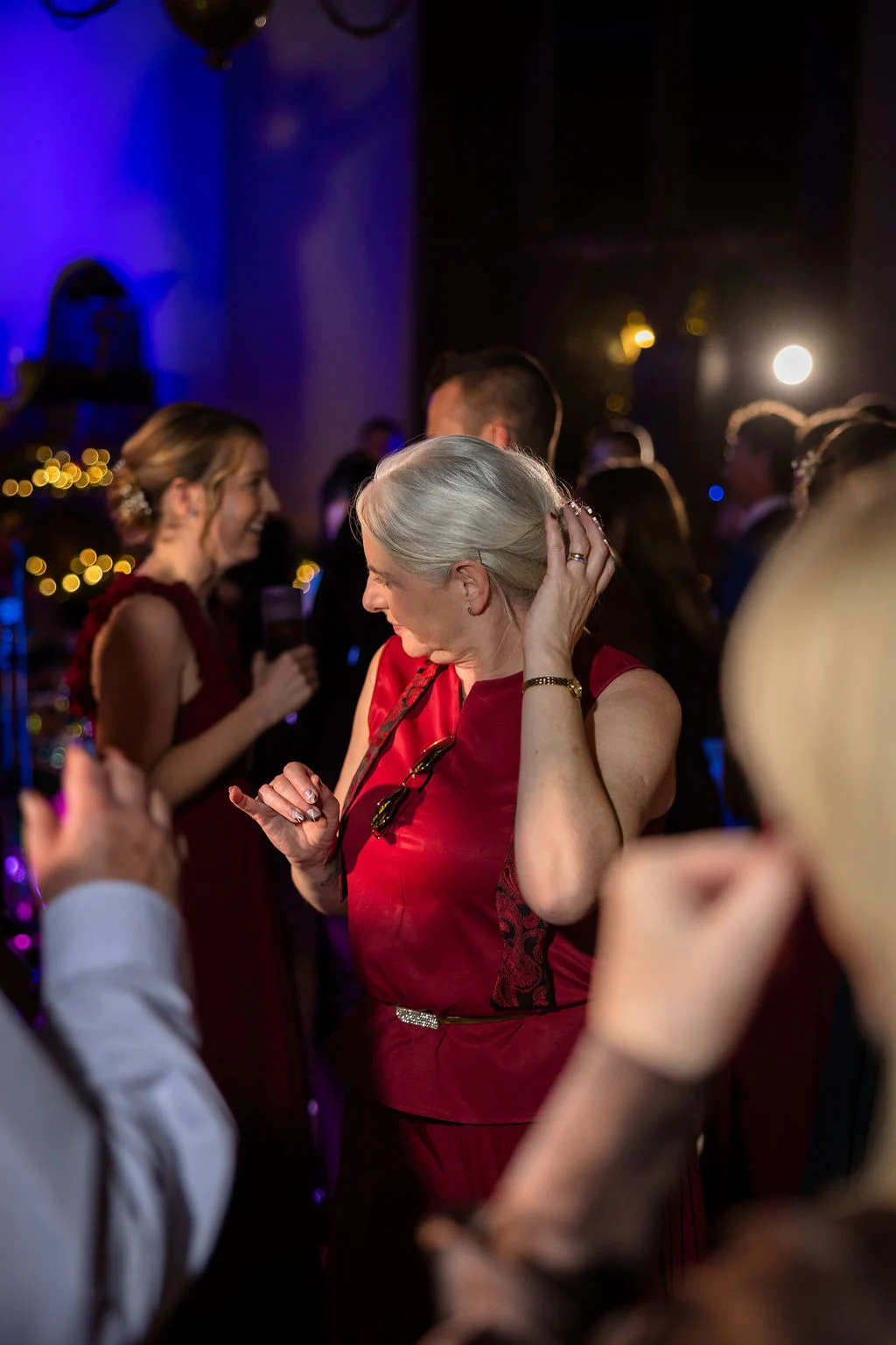 A mature guest with grey hair dances happily among the crowd during an evening wedding reception illuminated by ambient coloured lighting.