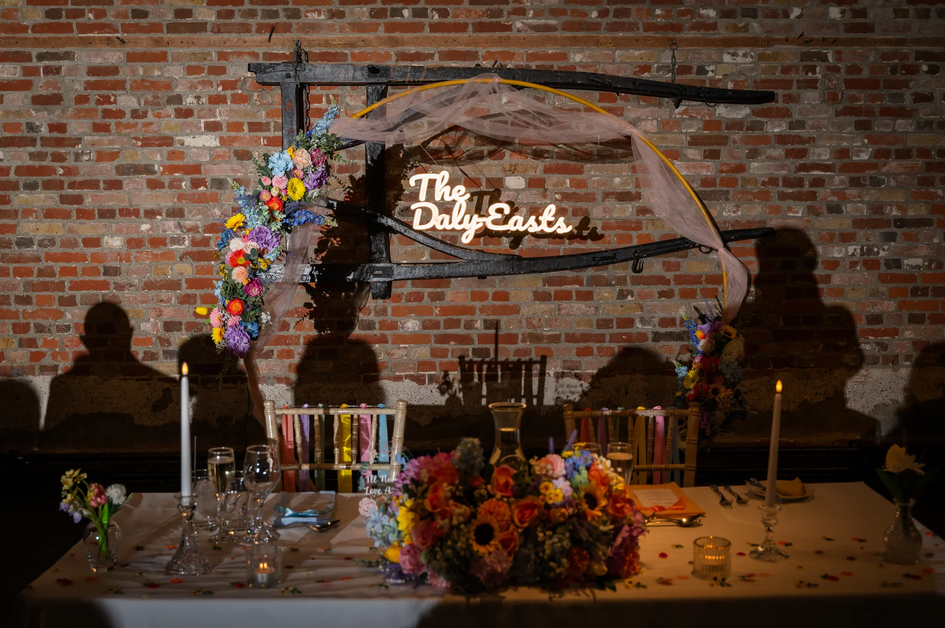 An intimate top table setup featuring a vibrant floral arrangement, taper candles, and a glowing custom neon sign against an exposed brick wall.