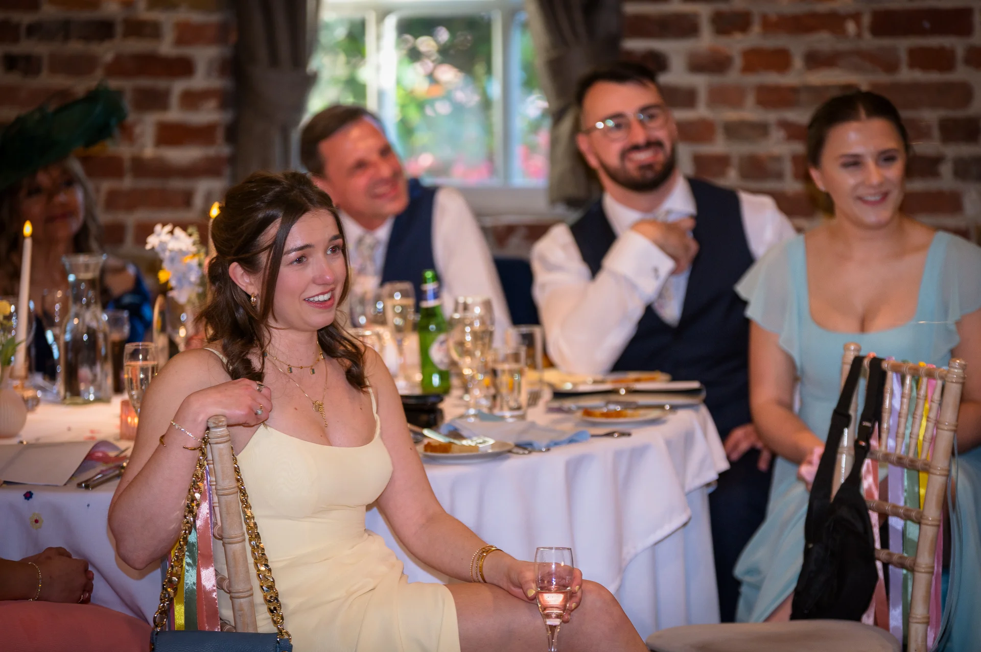 Wedding guests sit at a round dining table with a white tablecloth, laughing and smiling during the wedding speeches in a rustic room with exposed brick walls.