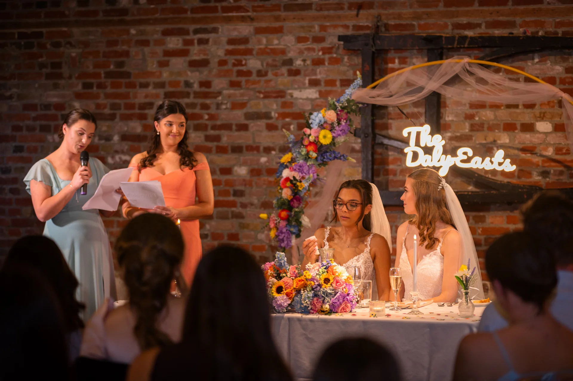 A bridesmaid gives a heartfelt speech at the top table while two brides listen attentively in front of a rustic exposed brick wall.