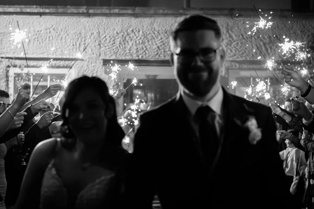 A newlywed couple walks through a tunnel of glowing sparklers held by cheering wedding guests at night.