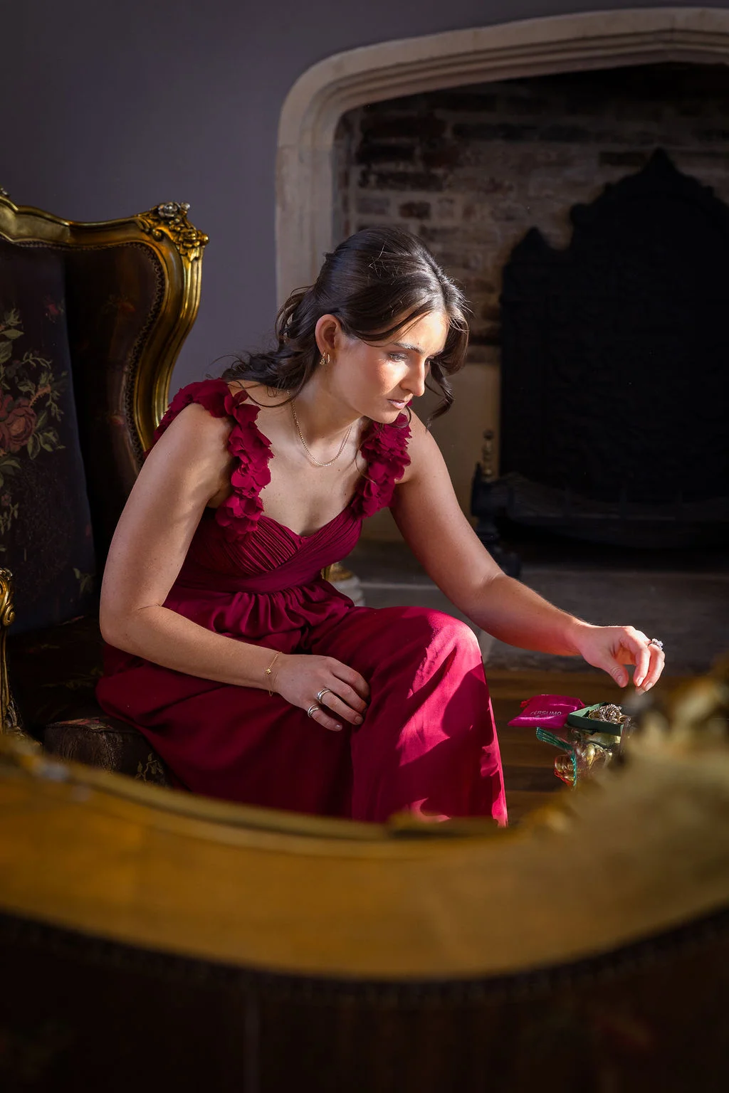 A bridesmaid in a ruby red gown and half-up hairstyle sits in an ornate armchair near a fireplace, reaching for jewellery during wedding preparations.