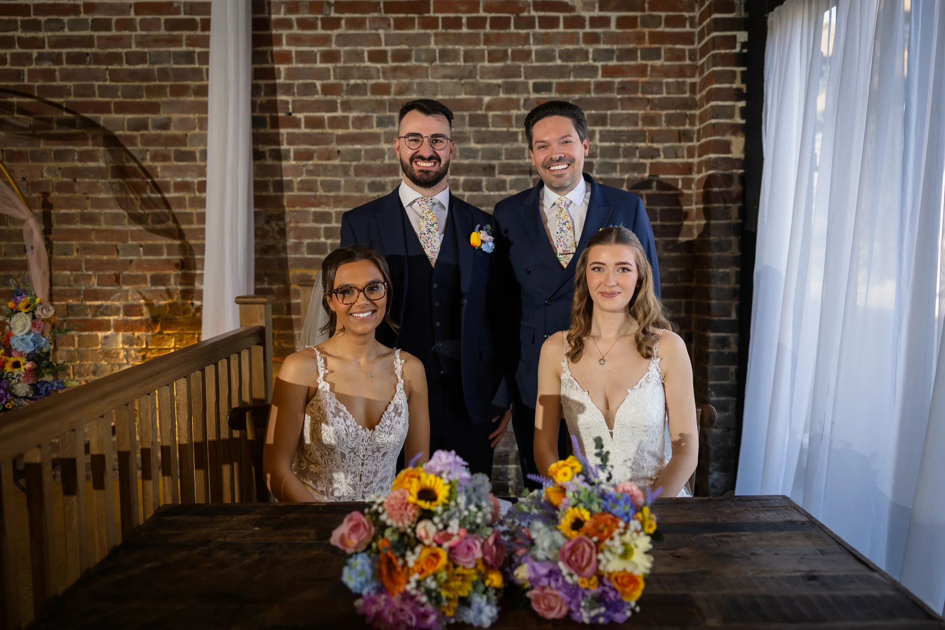 A newlywed couple and their groomsmen pose together smiling in front of an exposed brick wall behind a wooden table adorned with colourful floral bouquets.