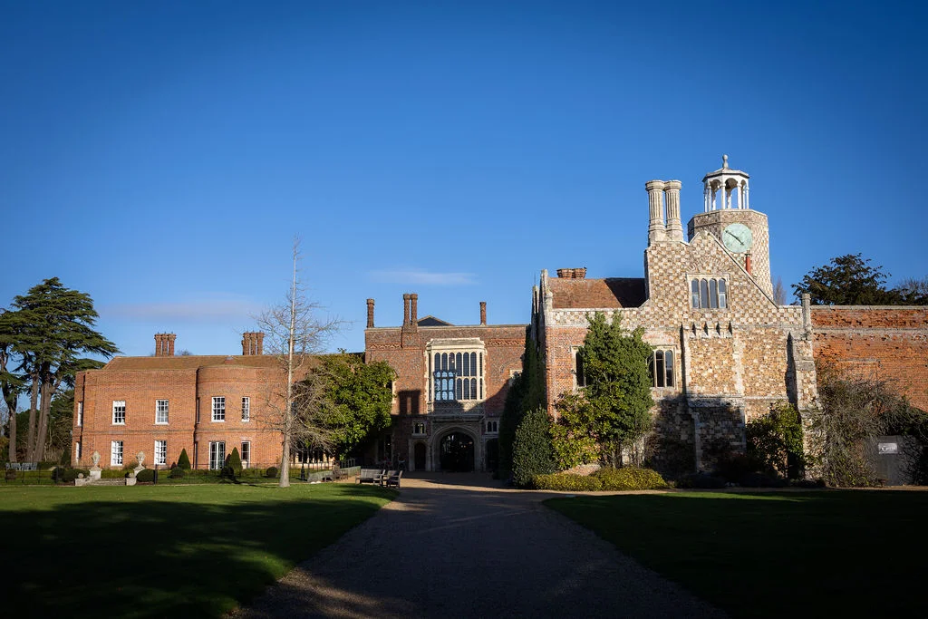 An impressive red brick stately home and manor wedding venue stands under a clear blue sky with a sweeping driveway leading to the entrance.