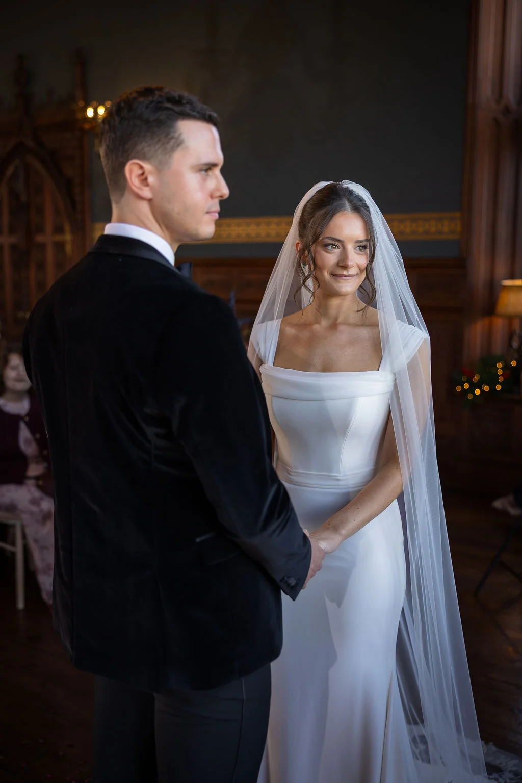 A newlywed couple holds hands during their ceremony inside a wood-panelled room, with the bride looking over towards the groom while wearing a classic square-neck gown and chapel-length veil.