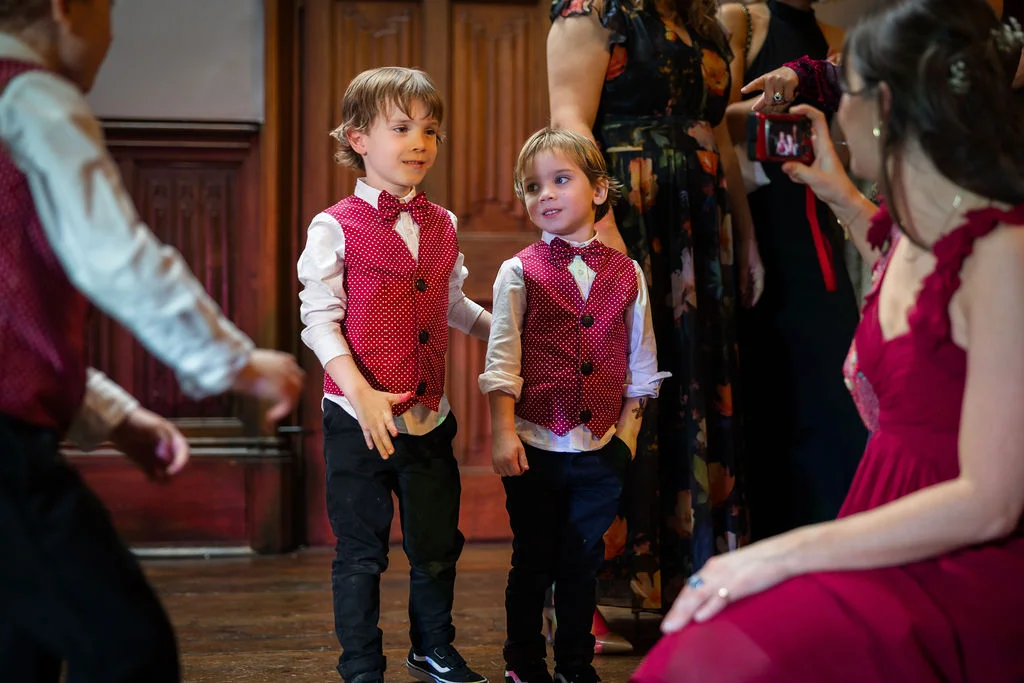 Two young page boys wearing matching red polka dot waistcoats and bow ties stand indoors on wooden flooring while guests look on.