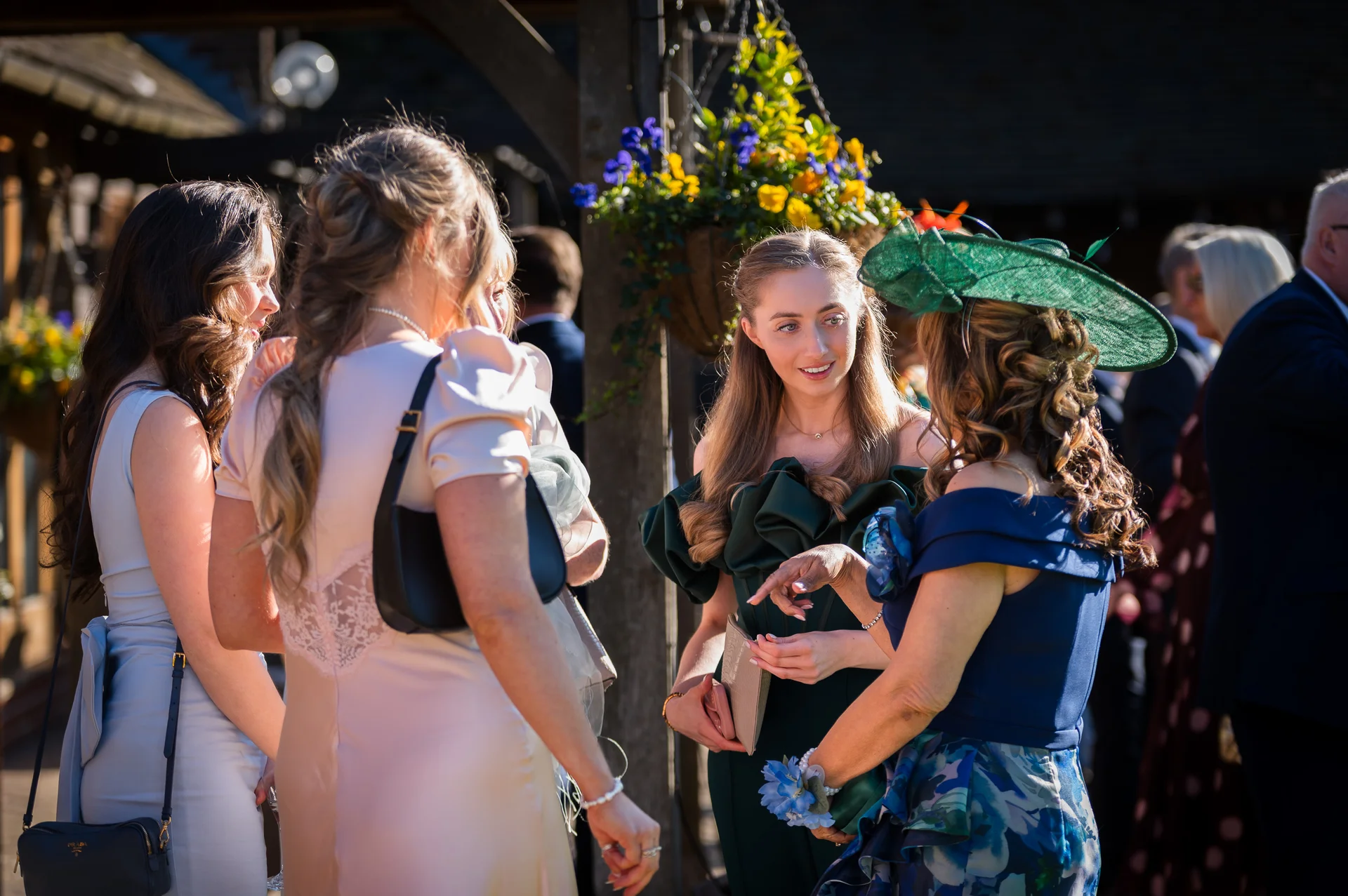A group of wedding guests mingle outdoors in the bright sunlight, with women wearing stylish dresses and fascinators in shades of green and blue.