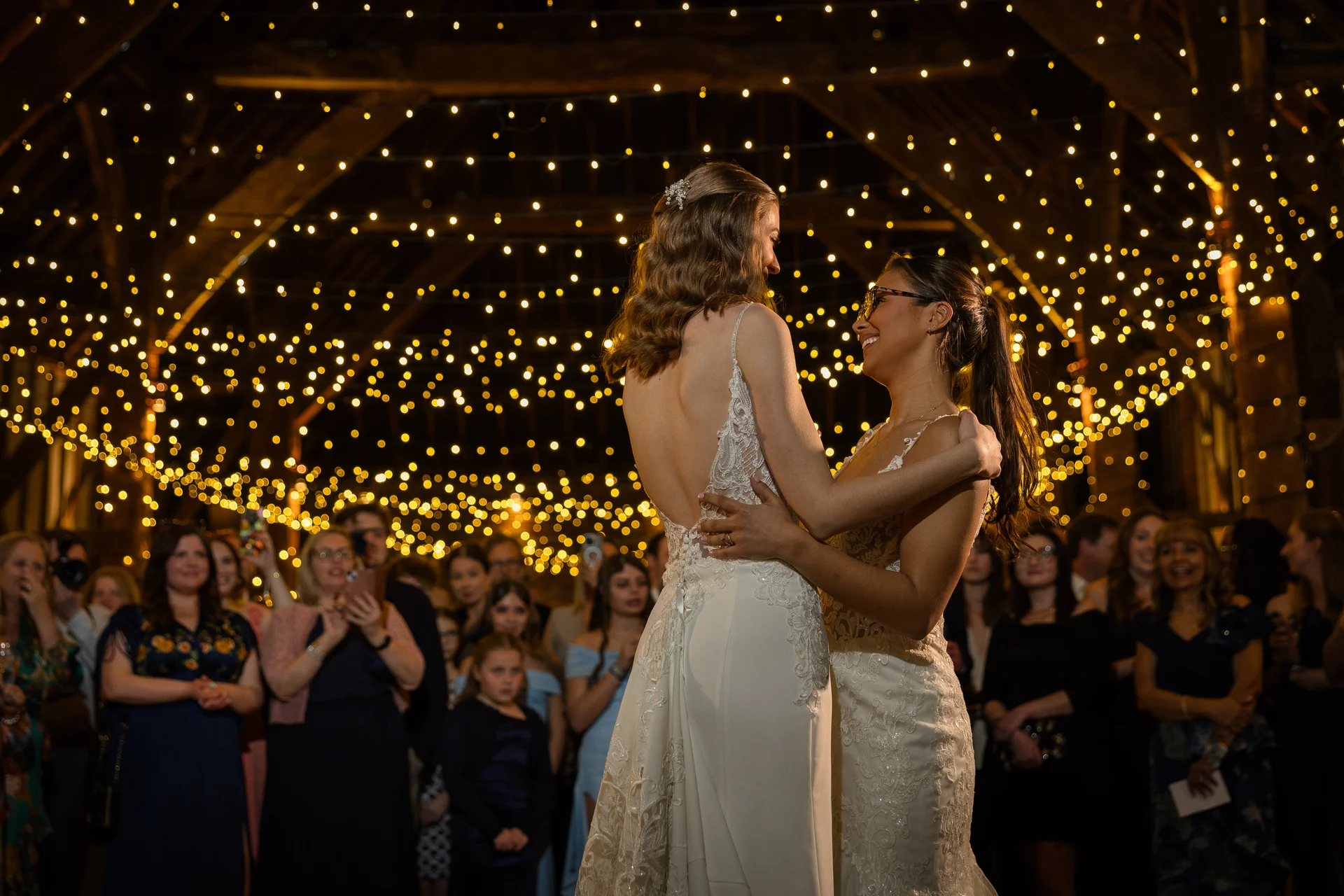 Two brides share their first dance in a rustic barn illuminated by a magical canopy of twinkling fairy lights.