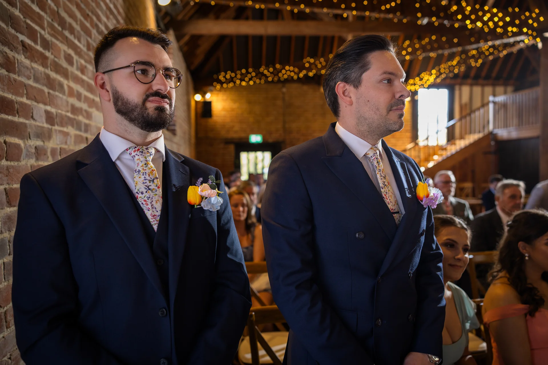Two grooms stand attentively side by side in a rustic brick barn adorned with fairy lights, both wearing navy suits and matching floral ties.