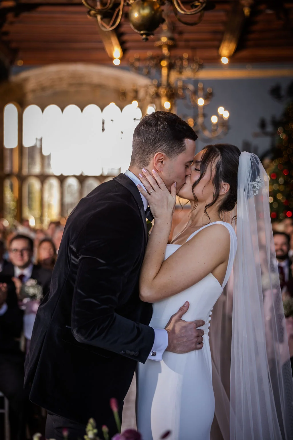 Newlyweds share their first kiss during an indoor ceremony at a manor house, with the groom wearing a velvet tuxedo and the bride in a sleek satin gown.