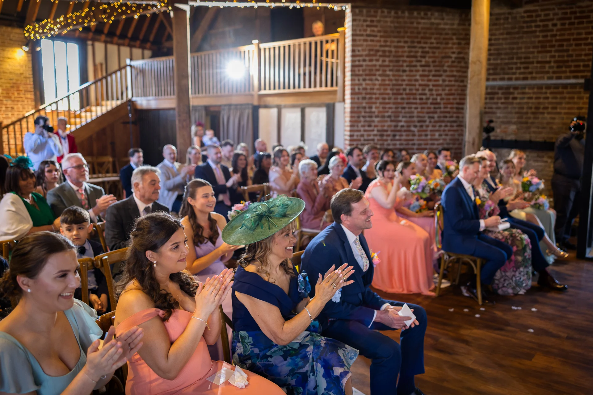 A joyful crowd of wedding guests applaud and smile inside a rustic brick barn venue with a mezzanine level and wooden staircase.