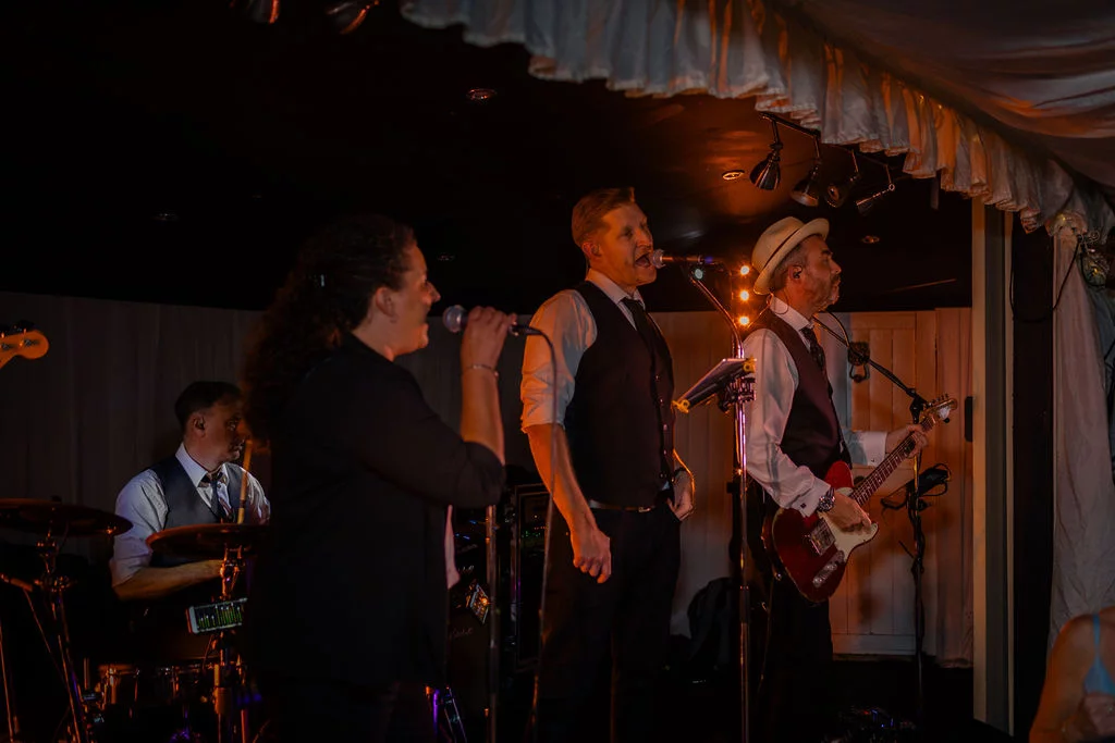 A lively wedding band performs on a stage under warm lighting inside a marquee during the evening reception.