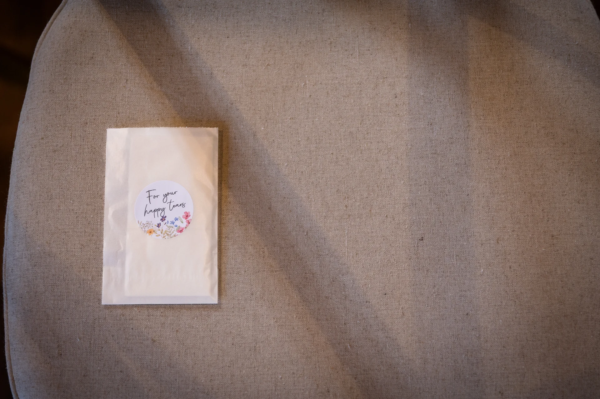 A close-up flat lay shot of a small paper packet labelled for happy tears resting on a textured neutral linen surface.