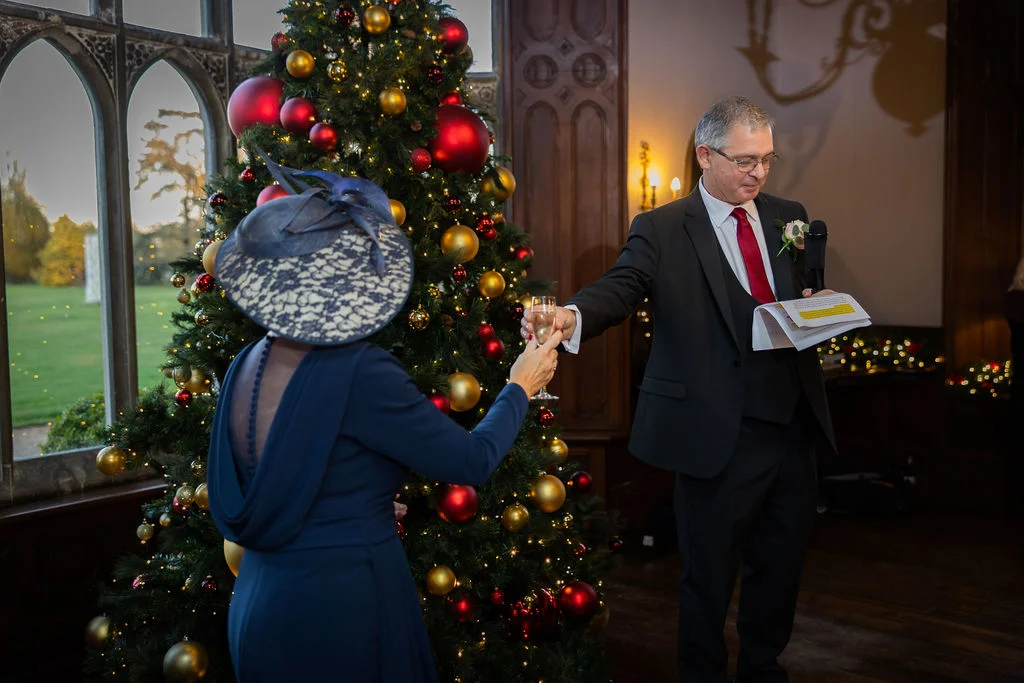 A guest in a navy dress and matching fascinator raises a glass to toast a speaker holding speech notes in front of a large Christmas tree.