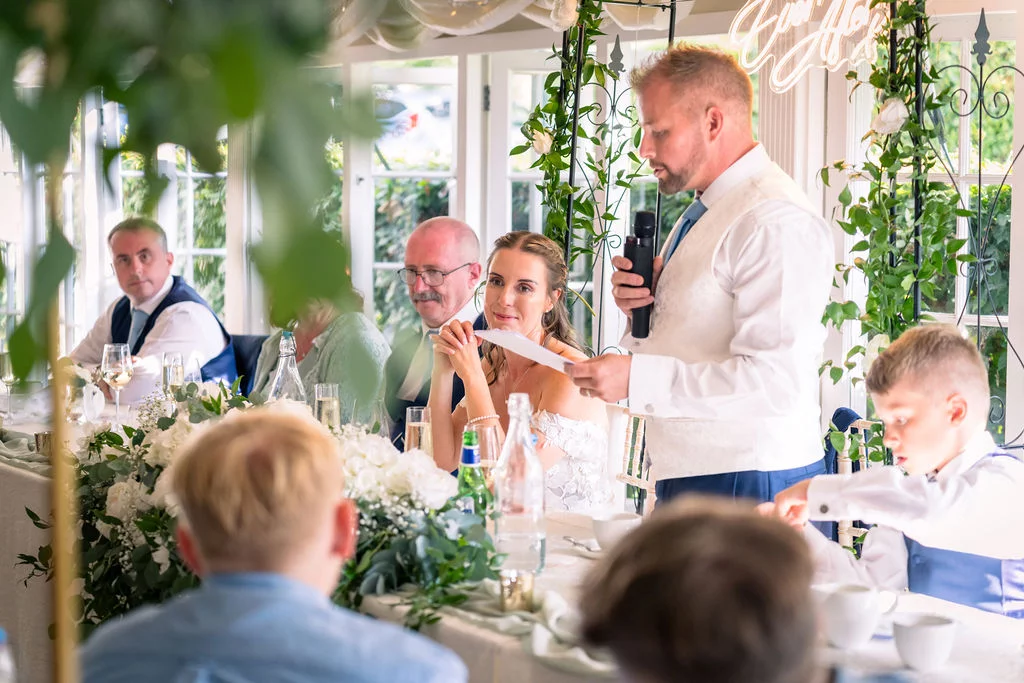 The groom stands delivering a speech while holding a microphone at the top table, with the smiling bride and wedding guests looking on in a bright conservatory venue.
