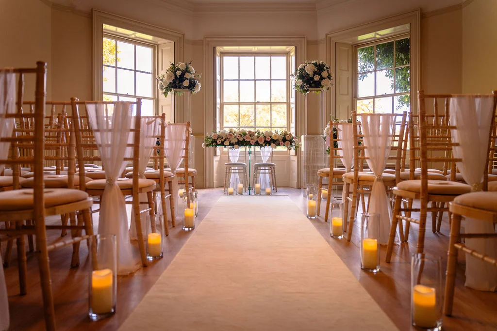 An elegant indoor wedding ceremony setup featuring a light-filled room with large sash windows, a cream aisle runner lined with glass cylinder candles, and chiavari chairs adorned with delicate fabric sashes.