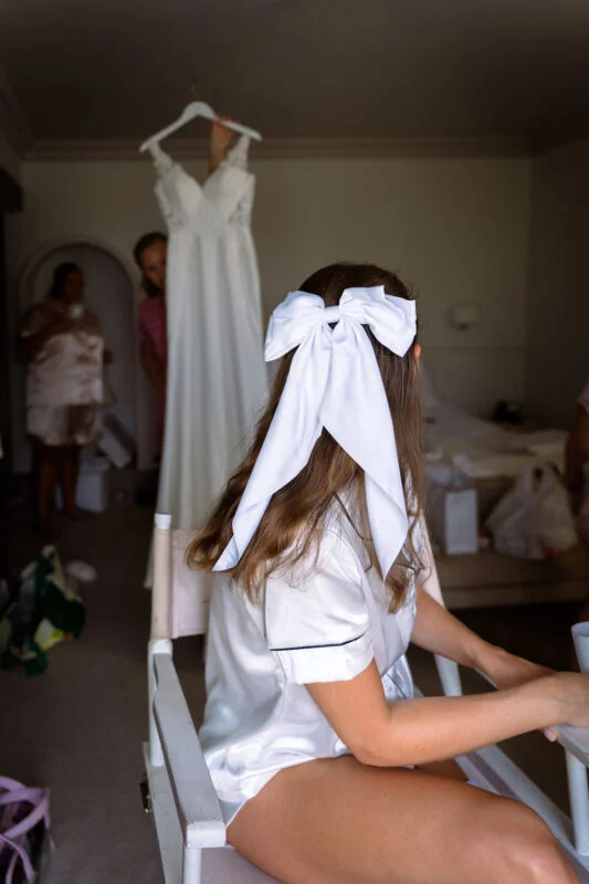 A bride sits in a chair during morning preparations wearing white silk pyjamas and a large white hair bow, while her wedding dress hangs in the background.
