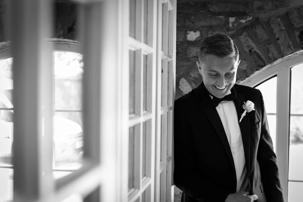 A smiling groom wearing a black tuxedo and bow tie looks down happily while standing near an exposed brick wall and window.