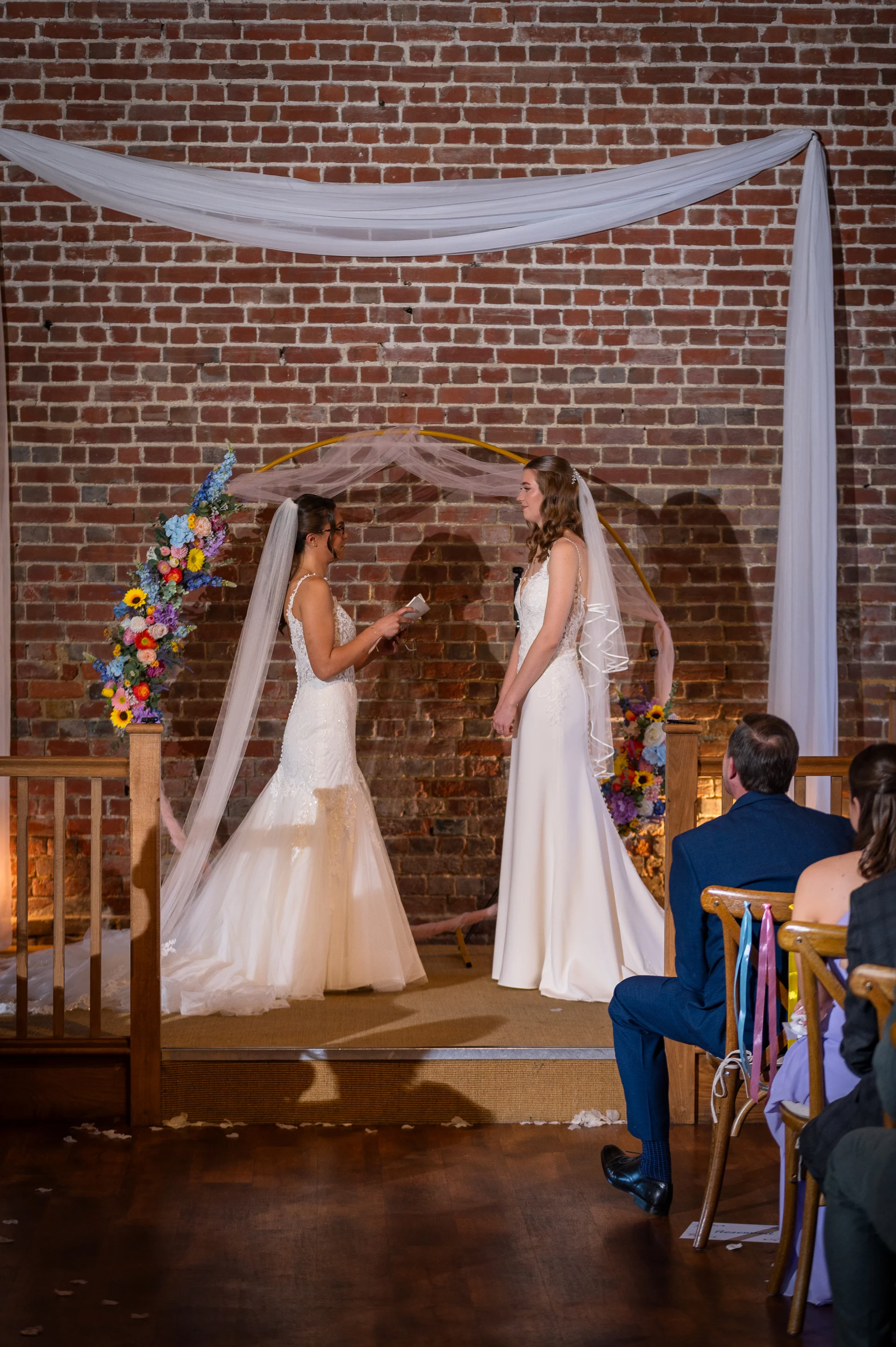 Two brides stand facing each other during their wedding ceremony in front of an exposed brick wall and a floral arch, while guests look on from the wooden seating.