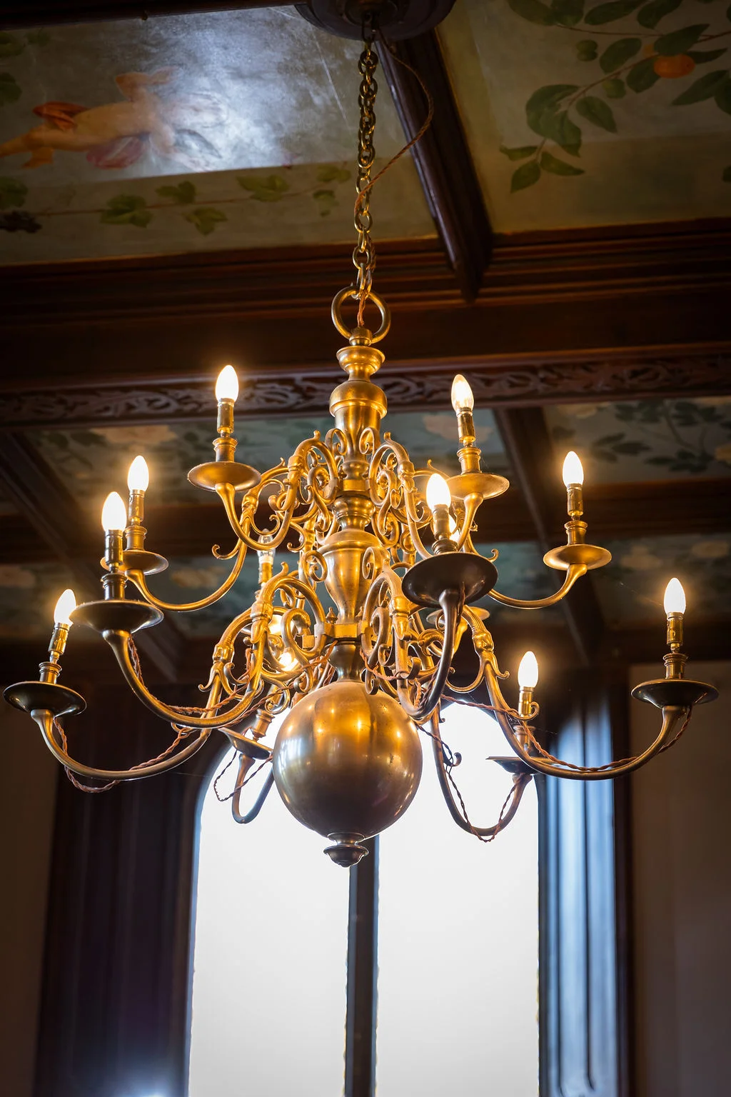 An ornate brass chandelier hangs from a painted ceiling with exposed wooden beams inside a historic wedding venue.