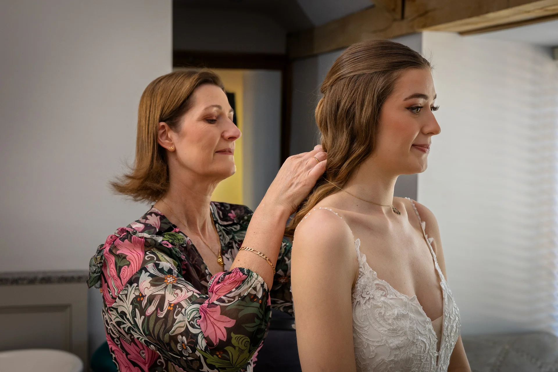 A mother gently arranges her daughter's hair in a half-up style while the bride wears a lace wedding dress during morning preparations.