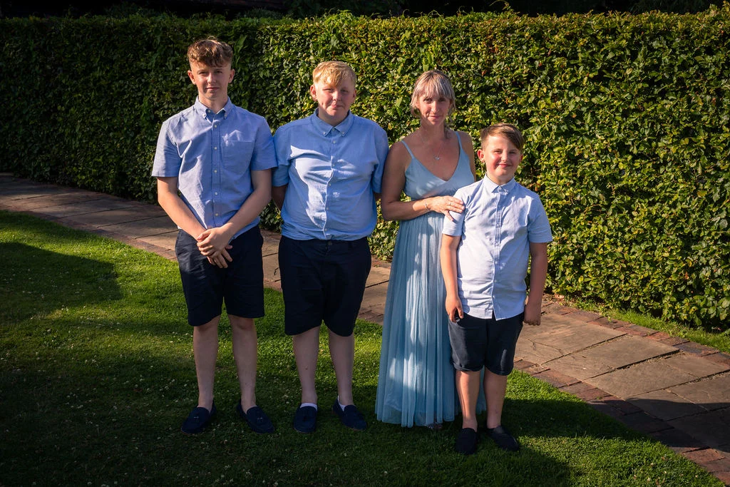 A woman in a blue dress posing outdoors in the golden sunlight alongside three boys wearing matching blue shirts and dark shorts.