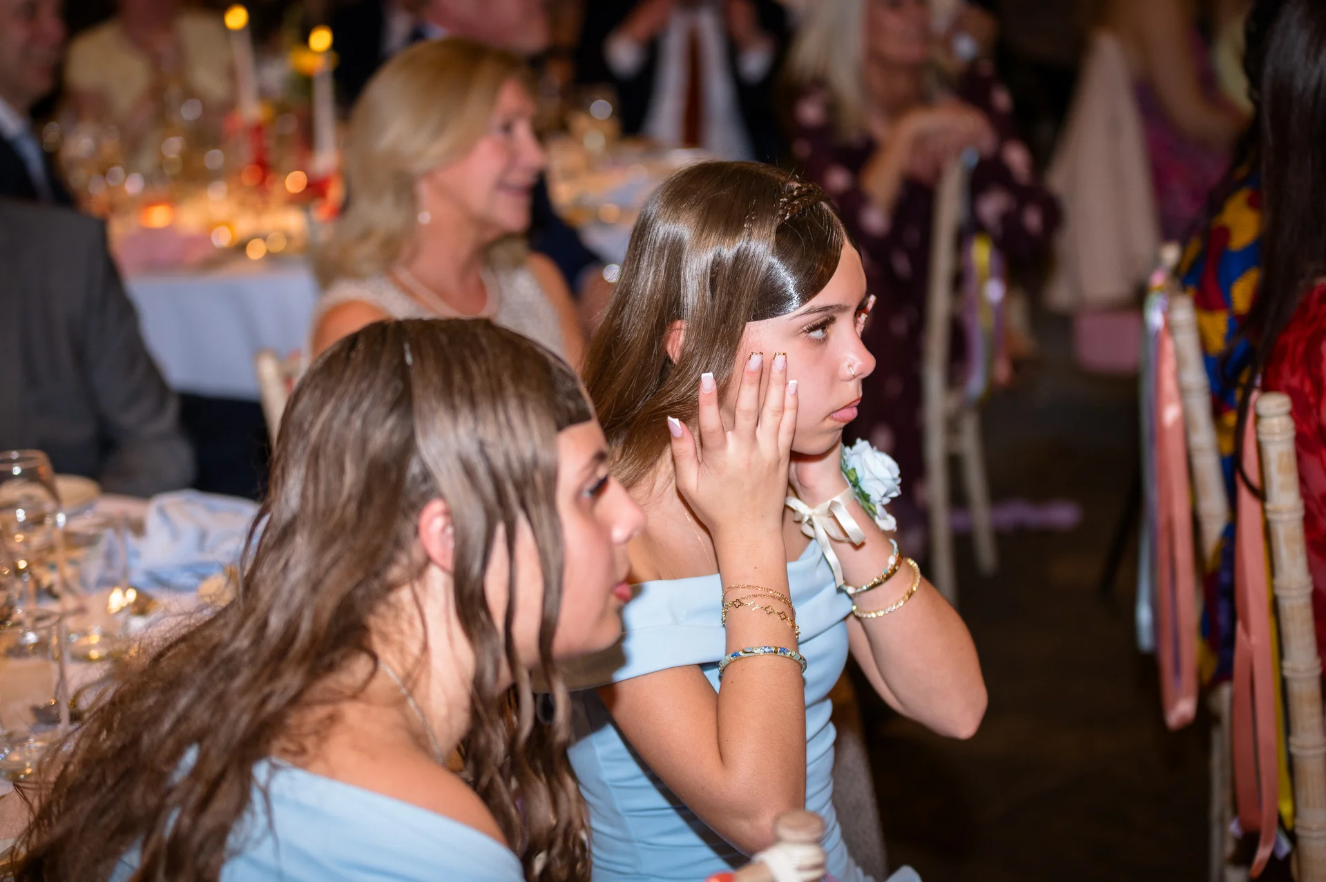 Two bridesmaids in matching dusty blue dresses sit at a reception table, watching intently and resting their hands gently on their faces during speeches.