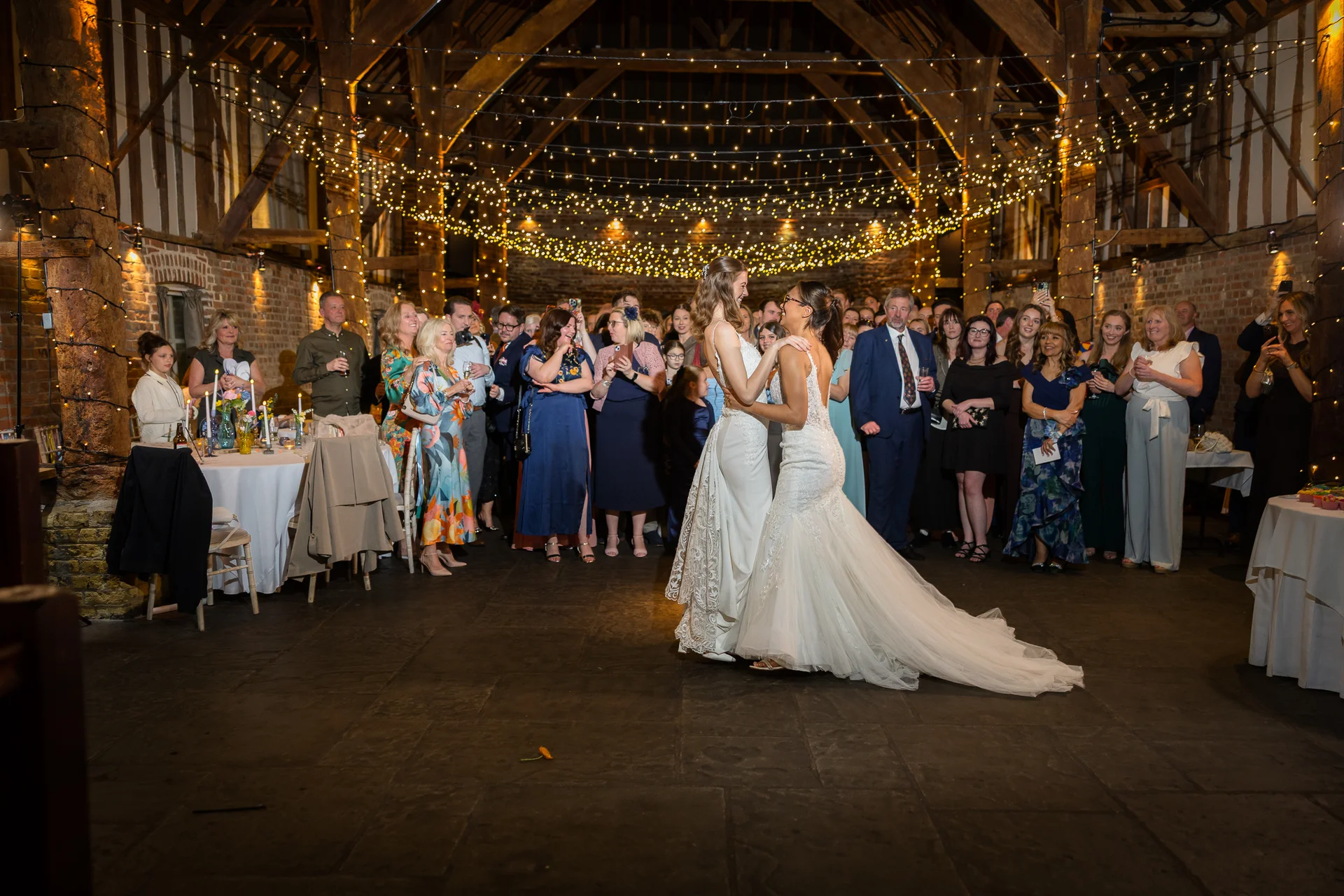 Two brides share their first dance in a stunning oak-framed barn illuminated by an extensive fairy light canopy while wedding guests watch with delight.
