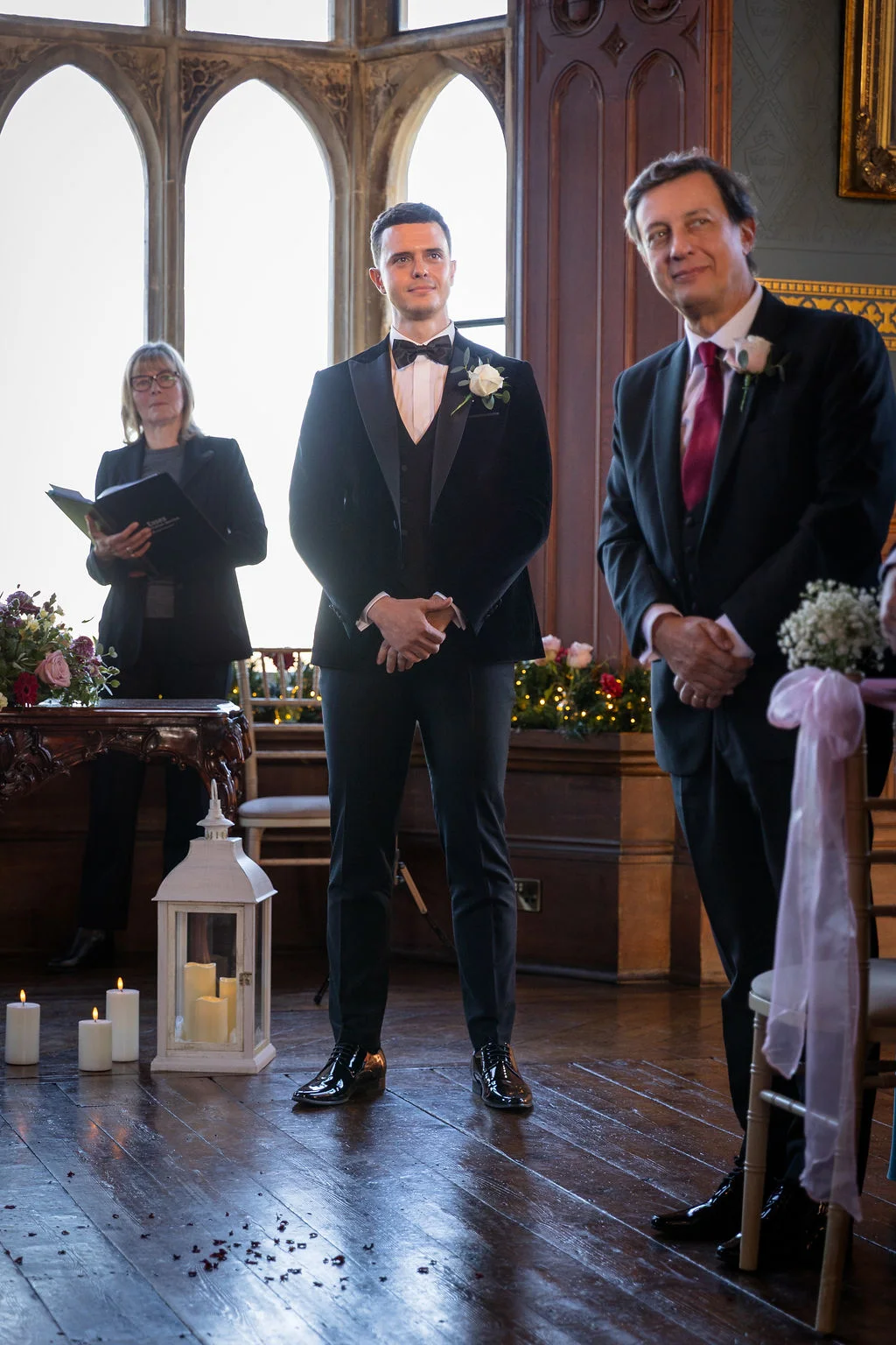 The groom stands attentively at the altar in a sharp black tuxedo with a bow tie, accompanied by another man and an officiant in a historic hall with large arched windows.