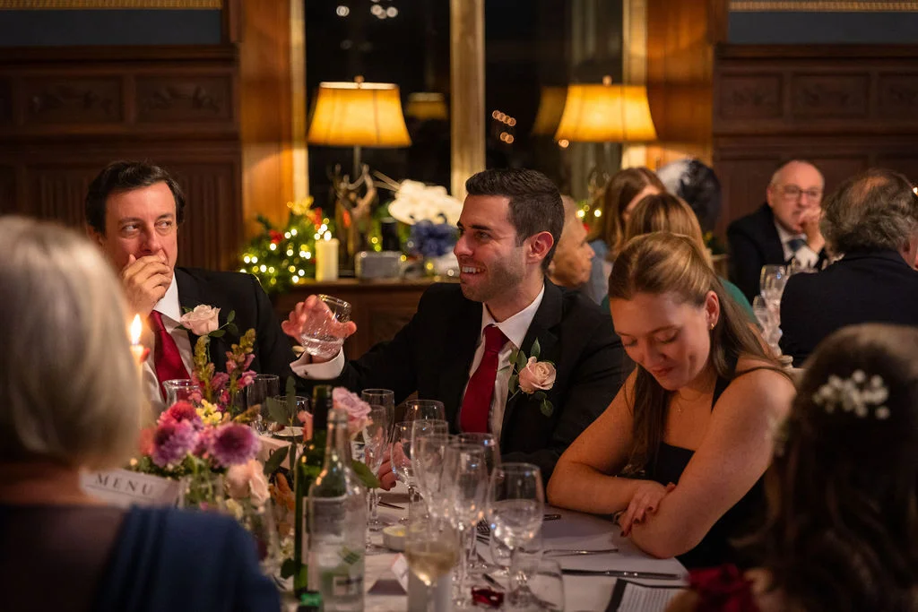 Wedding guests enjoy a candlelit wedding breakfast in a wood-panelled room while a man raises a glass and others laugh.