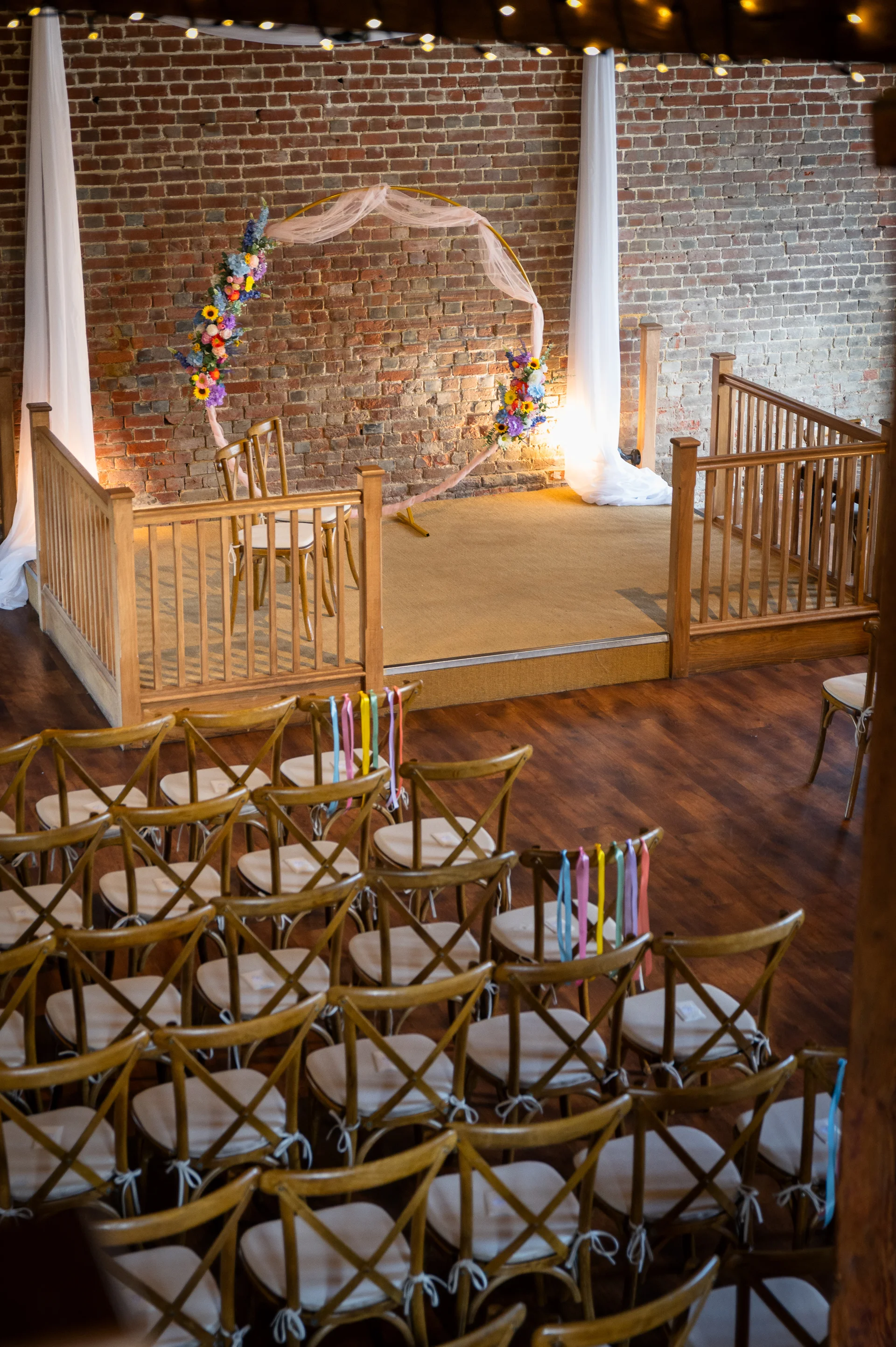 An empty wedding ceremony setup inside a rustic barn features a wooden stage with an exposed brick wall, a circular floral arch, and rows of wooden chairs adorned with colourful ribbons.