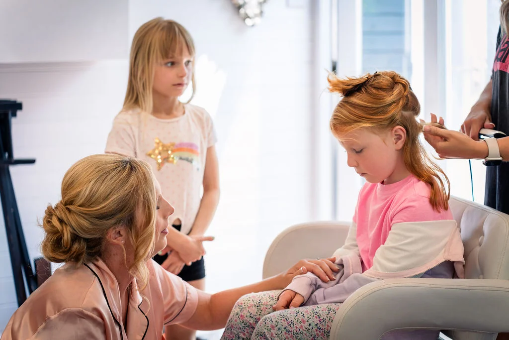 A young girl has her hair styled while a woman in a silk robe and another child look on during morning wedding preparations.