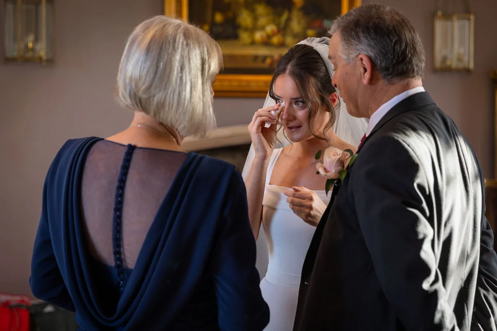 An emotional bride gently wipes away tears as she shares a tender, tearful moment with her parents indoors.