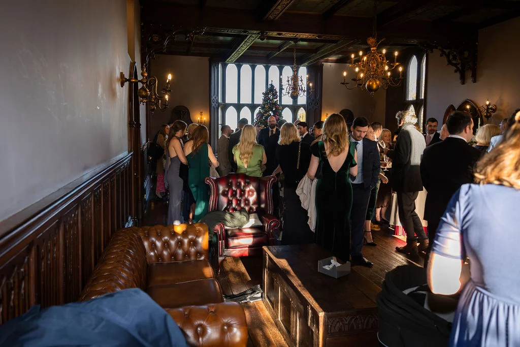 Guests mingle in a wood-panelled country house reception room featuring leather chesterfield sofas, vintage chandeliers, and warm winter afternoon sunlight streaming through tall windows.