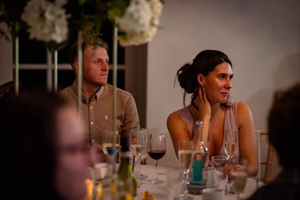 A guest and a groom are captured candidly during the wedding breakfast, sitting at a beautifully dressed table surrounded by wine glasses and tall floral centrepieces.
