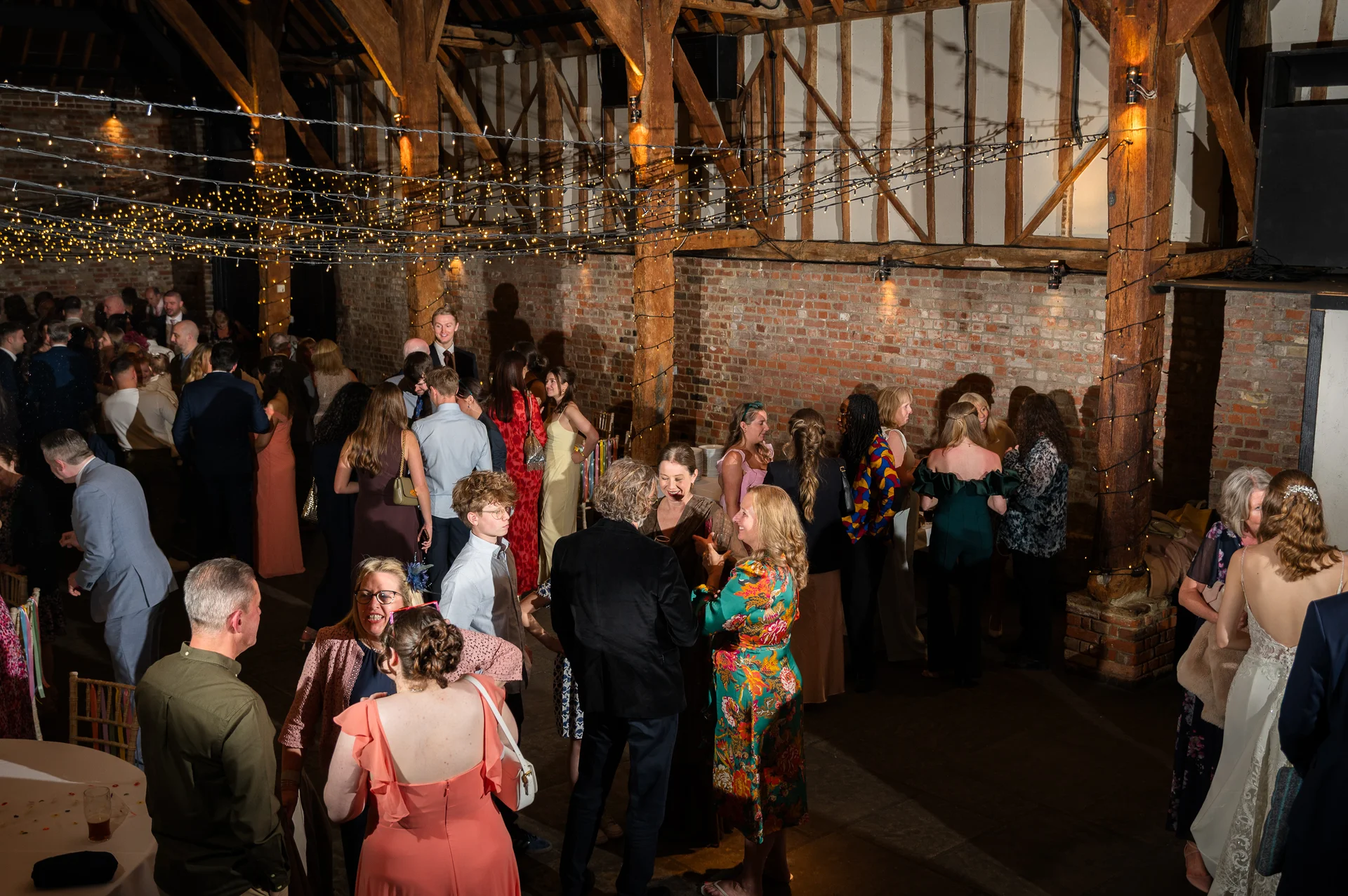 Wedding guests mingle during the evening reception inside a rustic oak-framed barn decorated with exposed brick walls and a canopy of fairy lights.