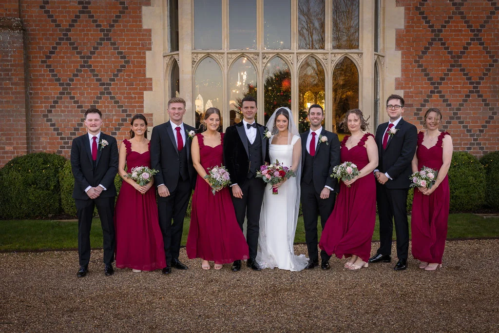 The bride, groom, and their wedding party stand in a neatly arranged group on a gravel path in front of a red brick country house with large arched windows.