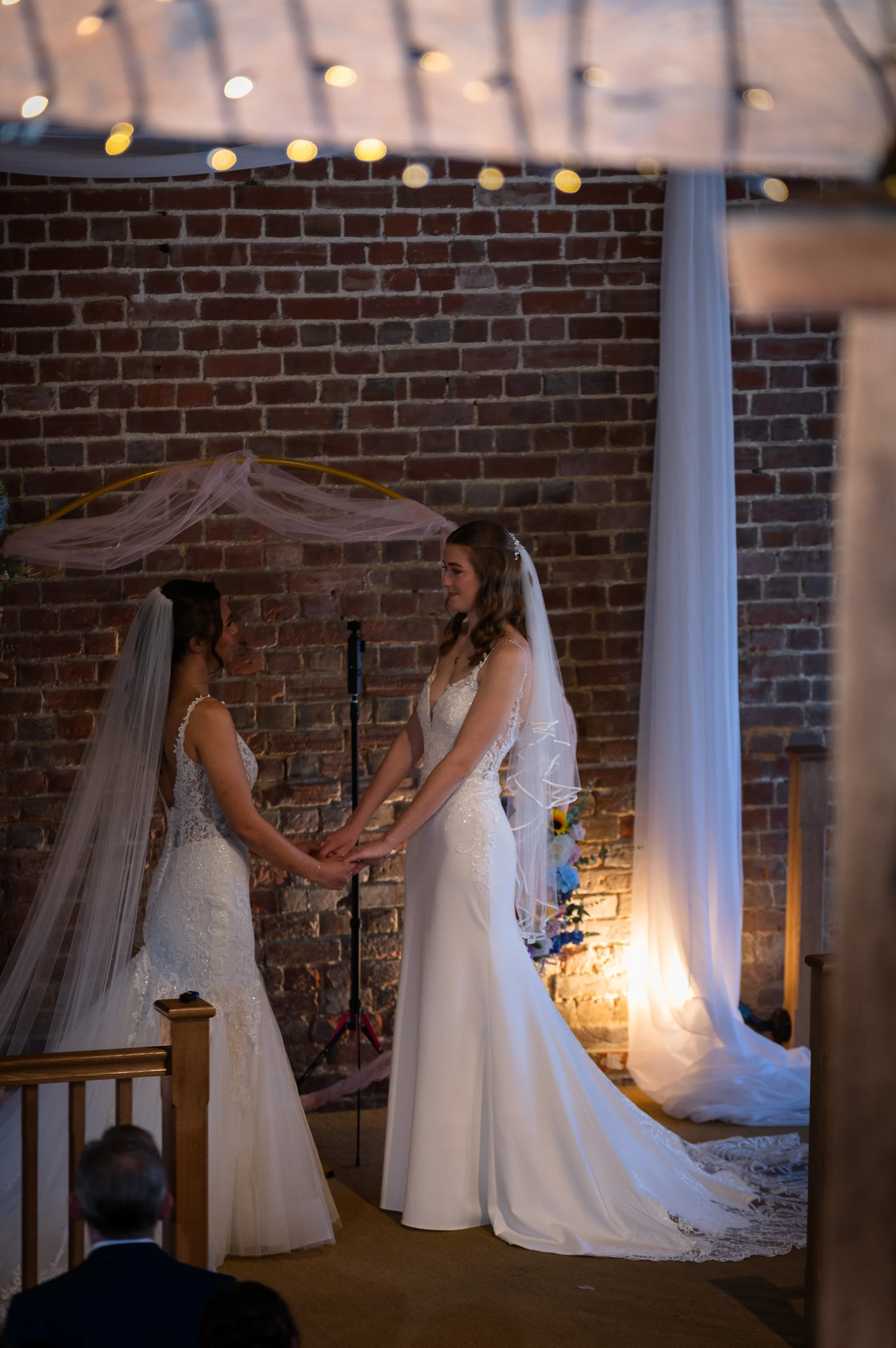Two brides stand hand in hand during their intimate wedding ceremony inside a rustic barn with exposed brick walls and twinkling fairy lights.