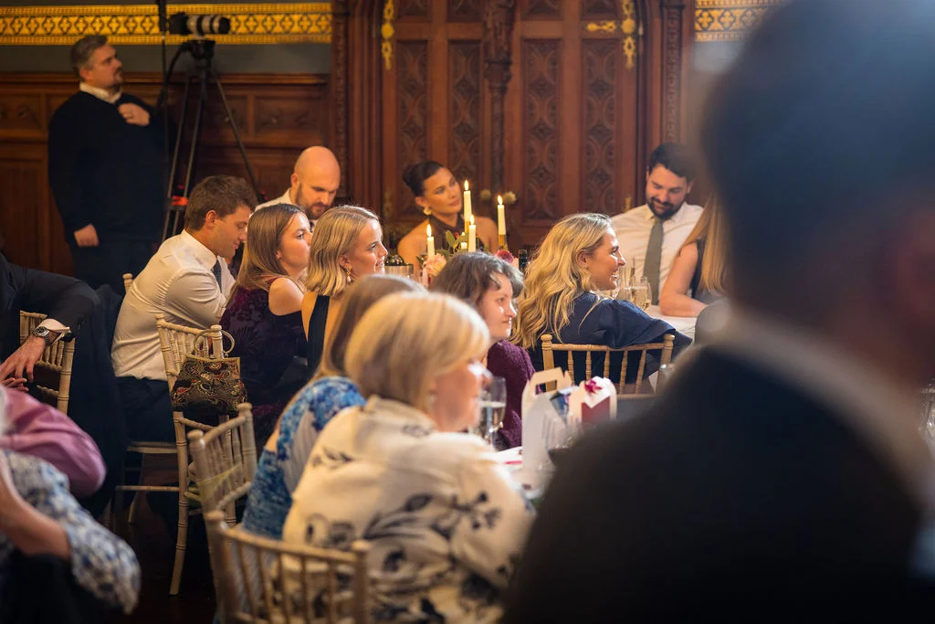 Wedding guests enjoy a lively wedding breakfast seated at a long table inside an elegant wood-panelled room illuminated by candlelight.