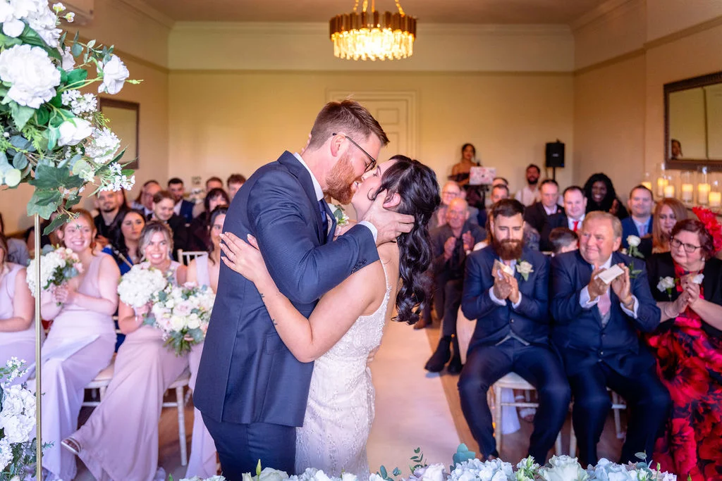 The bride and groom share their first kiss as a married couple at the front of a light-filled ceremony room while guests clap and cheer.