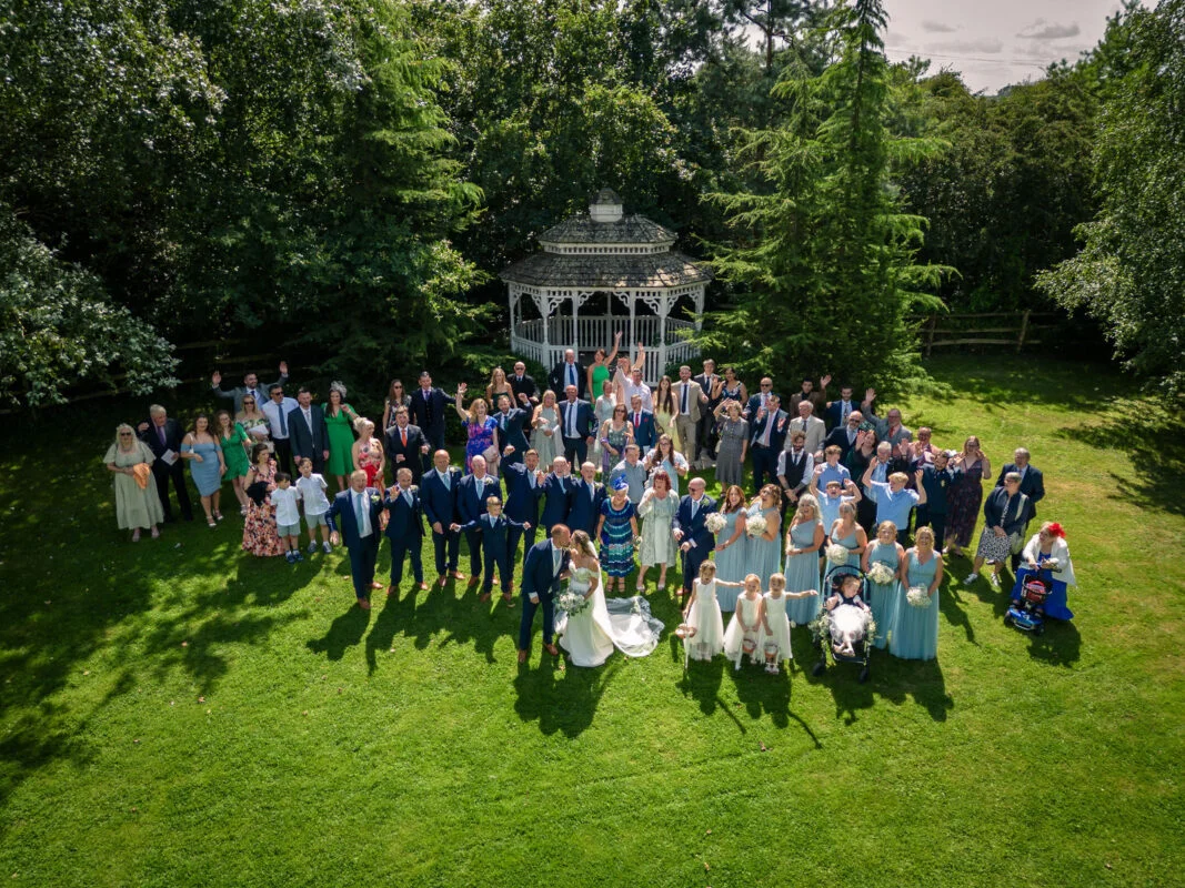 An aerial drone shot captures a large wedding party and guests gathered on a lush green lawn in front of a white gazebo on a sunny summer day.