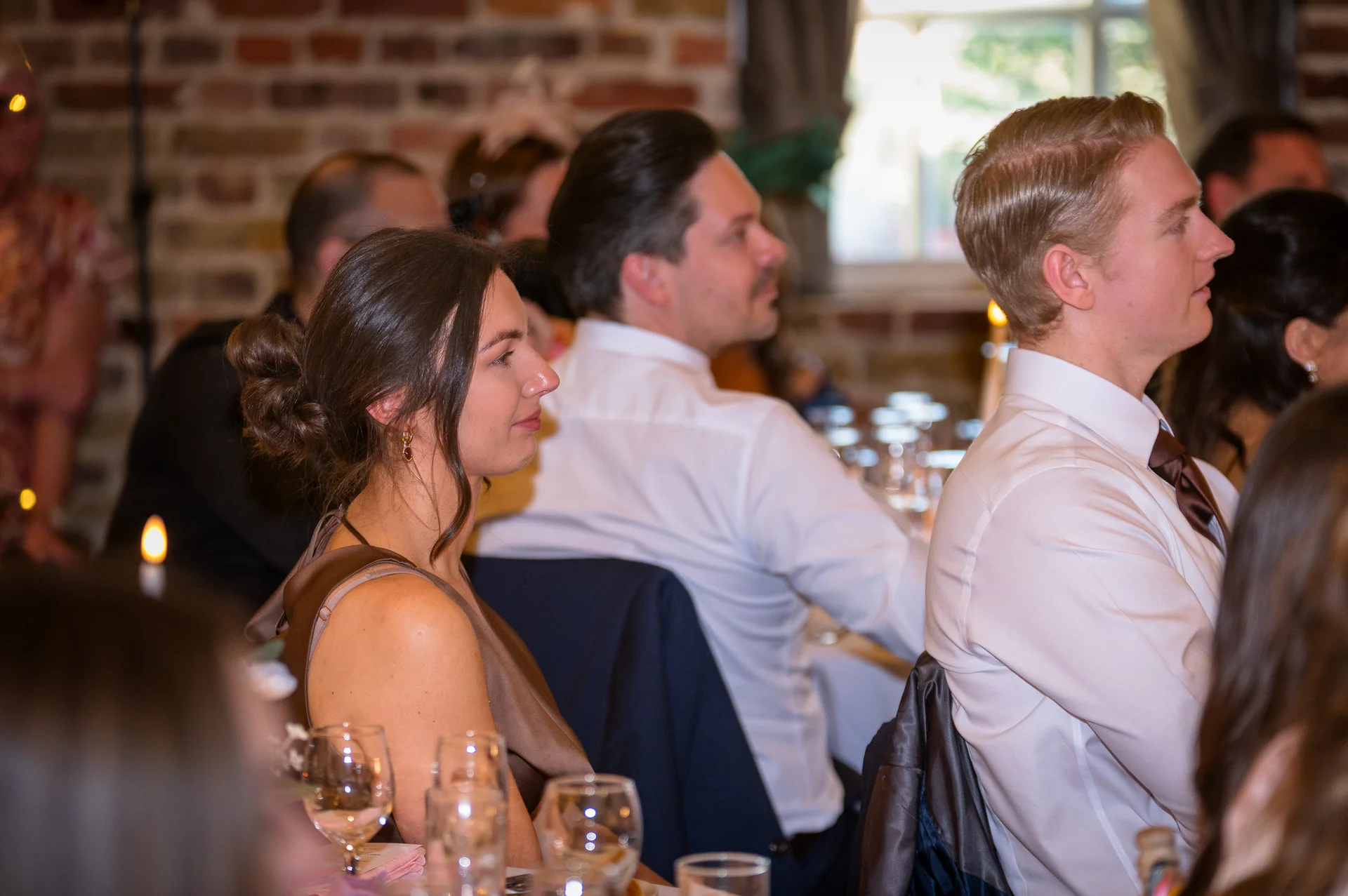 Wedding guests seated at tables during the reception speeches, listening attentively with warm indoor lighting and an exposed brick background.