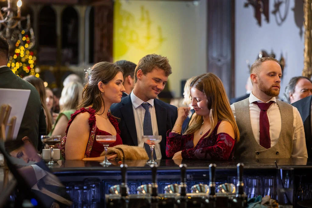 Wedding guests in stylish attire enjoy drinks and conversation gathered around a dimly lit indoor bar during the reception.
