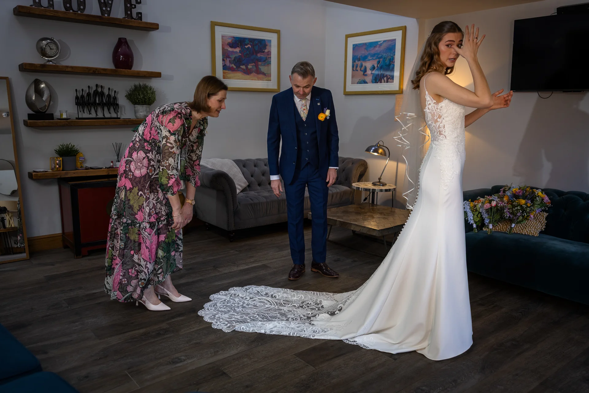 A bride checks her reflection in a mirror as her mother inspects the lace train of her gown and her father looks on proudly in a blue suit.