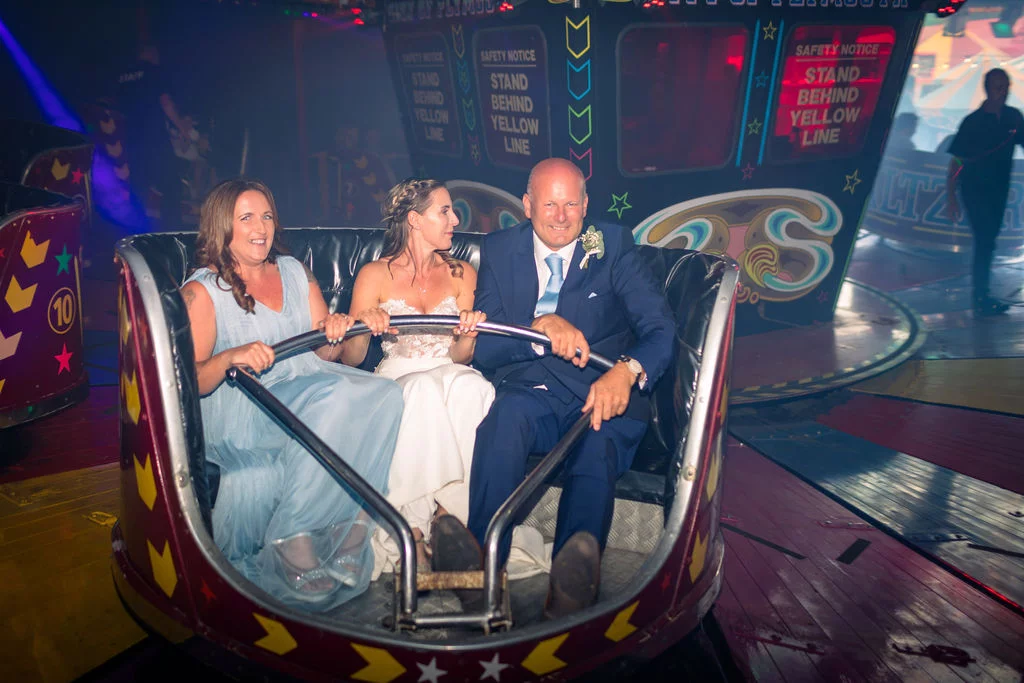 A bride, groom, and guest laugh together while riding a fairground attraction during an evening wedding reception.