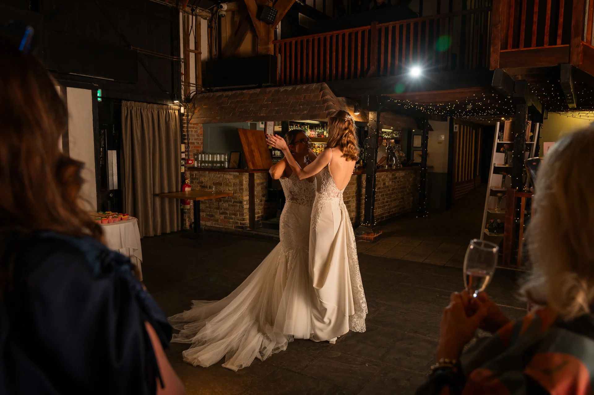 Two brides share their first dance in a rustic barn venue illuminated by string lights, surrounded by guests watching from the foreground.