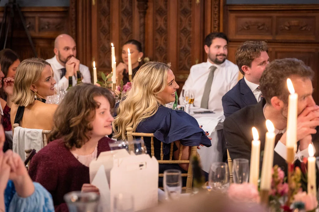 Wedding guests sit at a candlelit banquet table inside a wood-panelled room, laughing and listening attentively during the wedding breakfast speeches.