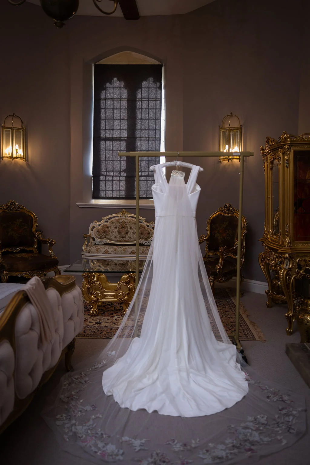 A stunning white wedding dress with a dramatic embroidered cathedral veil hangs on a rack inside an opulent, historically decorated room lit by natural window light.