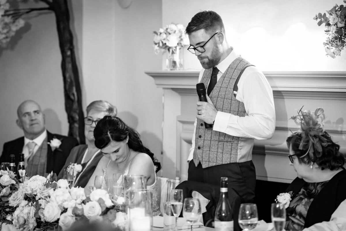 A groom wearing a tweed waistcoat and glasses delivers a speech holding a microphone during the wedding reception.
